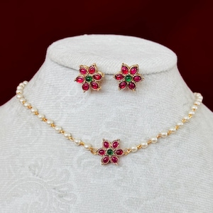 May include: A gold-toned necklace and matching stud earrings set. The necklace features a central flower pendant and is strung with small pearls. The earrings and pendant have red and green gemstone accents in a floral design.