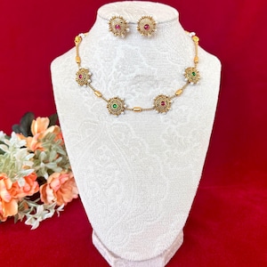 May include: A gold necklace and earring set displayed on a white mannequin. The necklace has circular pendants with red, green, and white accents. The earrings have matching red accents. The jewellery is set against a red background.