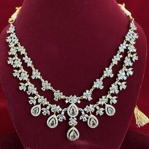 Pu&ograve; includere: Una collana a due fili con un design floreale. La collana presenta pietre chiare e scintillanti incastonate in un metallo color oro. Ciondoli a forma di goccia pendono dal filo inferiore. La collana &egrave; esposta su uno sfondo di velluto rosso.