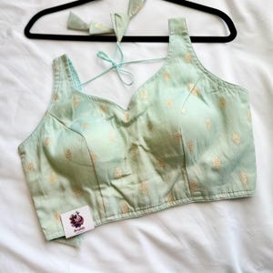 May include: A light green blouse with a V-neck and gold floral pattern. The blouse is hanging on a black hanger. The blouse has a tag that says "Anafab".