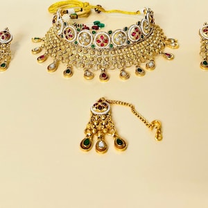 May include: A gold-toned jewelry set featuring a choker necklace, earrings, and a headpiece. The necklace and headpiece have a central design with red, green, and white accents. The earrings and necklace have teardrop-shaped pendants.