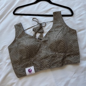 May include: A taupe-colored sleeveless top with a V-neckline, embellished with small, shimmering details. The top is hanging on a black hanger against a white background. A small label with a purple flower logo is visible.