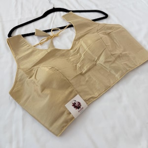 May include: A sleeveless gold-colored blouse with a V-neckline and a tie-back closure. The garment features a subtle sheen and a small white square tag on the lower front with a graphic logo and the text AnaFab printed in dark lettering.