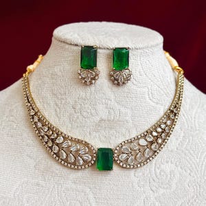 May include: A gold-toned necklace and earring set. The necklace features a large emerald-green rectangular stone at the center, with a curved design adorned with clear, teardrop-shaped stones. Matching emerald-green rectangular earrings complete the set.