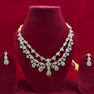 Pu&ograve; includere: Un set di collana e orecchini dorati, esposto su un busto di velluto rosso. La collana presenta un design floreale con pietre chiare e scintillanti e pendenti a goccia. Gli orecchini rispecchiano il design.