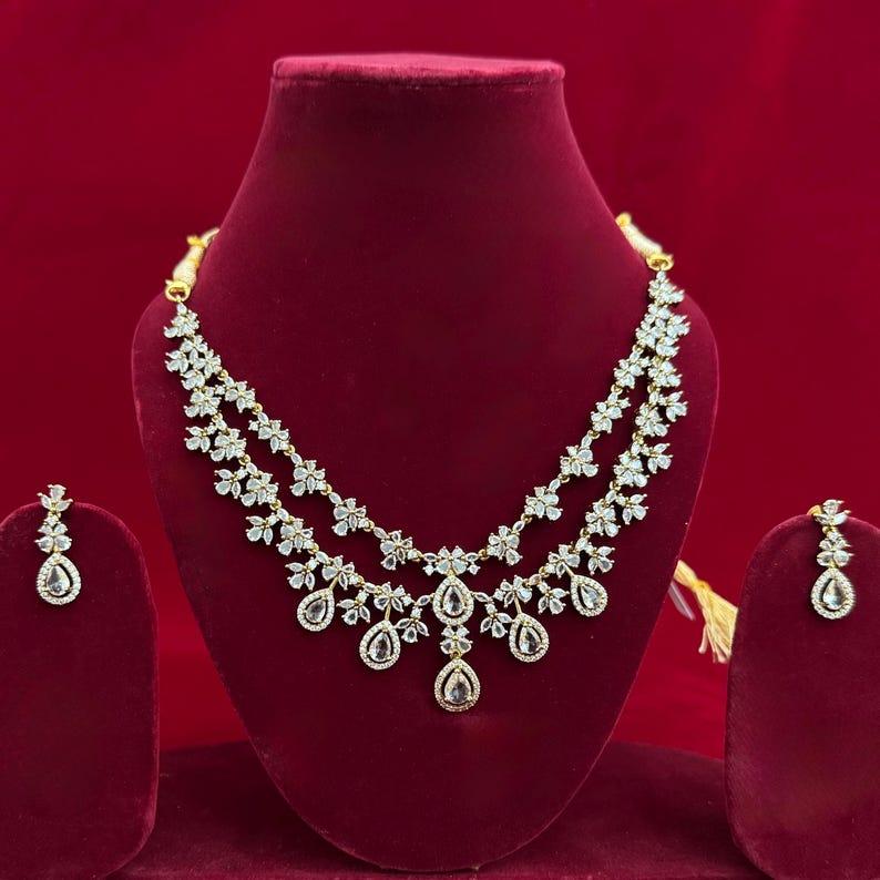 Pu&ograve; includere: Un set di collana e orecchini dorati, esposto su un busto di velluto rosso. La collana presenta un design floreale con pietre chiare e scintillanti e pendenti a goccia. Gli orecchini rispecchiano il design.