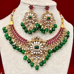May include: A gold-toned necklace and matching earring set featuring a central pendant with a rectangular green stone, surrounded by white crystals, red leaf-shaped accents, and dangling green beads with small white pearls throughout the design.