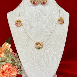 May include: A gold-toned jewelry set featuring a necklace and matching earrings. The necklace has a pendant with a green gemstone, and the earrings and necklace pendants have a crescent moon design with red gemstones. The set is displayed on a white bust.