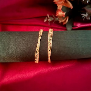 May include: Two gold bangles with intricate openwork designs. The bangles are adorned with small, colorful gemstones. They rest on a dark green surface against a backdrop of red fabric and flowers.