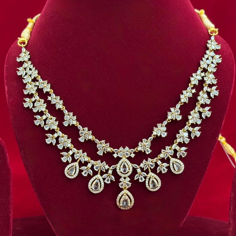 Pu&ograve; includere: Una collana dorata con due fili di accenti a forma di fiore e pendenti a goccia. La collana &egrave; adornata con pietre chiare e scintillanti, creando un look glamour. Il gioiello &egrave; esposto su una superficie di velluto rosso.