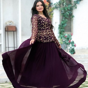 Könnte beinhalten: Eine Frau in einem dunkelvioletten Abendkleid. Das Kleid hat ein V-Ausschnitt-Oberteil mit goldenen Blumenstickereien und langen Ärmeln. Der Rock ist ein fließendes, plissiertes Design. Eine goldene Halskette ist sichtbar.
