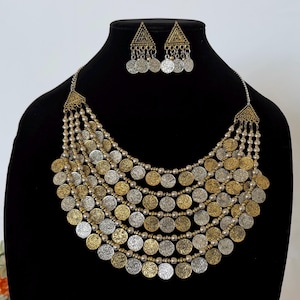 May include: A multi-layered necklace and matching triangular earring set featuring alternating gold and silver-toned circular coin charms suspended from metallic chains, accented with small round beads throughout the layered design.