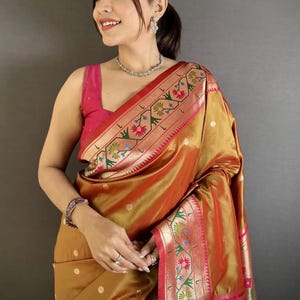 May include: A woman wearing a gold and red saree with floral embroidery. The saree has a pink border and is draped over a pink top. She is also wearing a silver necklace and earrings.