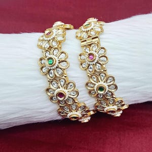 May include: Two gold-colored bangles with a floral design. Each flower has clear, sparkling accents and a colored center stone. The bangles are resting on a white, fluffy surface.