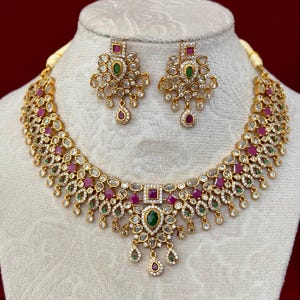 May include: A gold-toned necklace and matching earring set featuring clear white stones, square-cut pink stones, and oval-shaped green stones. The necklace has a central pendant design with dangling elements, and the earrings mirror this intricate pattern.