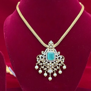 May include: A gold-toned necklace with an ornate pendant featuring a square, emerald-green gemstone at its center. The pendant is embellished with clear crystals and small, dangling pearls. The necklace is displayed on a red surface.