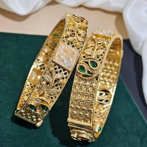 May include: Two gold bangles with intricate floral and geometric patterns. The bangles feature green and red gemstones, and a square, off-white inlay. The jewelry is displayed on a dark green surface with a white fabric in the background.