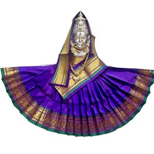 Puede incluir: Una estatua de deidad plateada con un rostro detallado, adornada con un collar y un tocado dorados. La estatua está envuelta en un vibrante sari púrpura con bordes dorados y verdes. El sari está elegantemente drapeado, creando un efecto fluido.