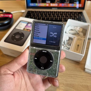Könnte beinhalten: Ein iPod mit transparentem Gehäuse, das die internen Komponenten zeigt, mit einem schwarzen Steuerkreuz und einem Bildschirm, auf dem das iPod-Menü angezeigt wird. Der Bildschirm zeigt Optionen wie Musik, Videos und Fotos an. Das Gerät wird in einer Hand gehalten, mit einer Box und einem Laptop im Hintergrund.