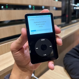 May include: A black iPod Classic music player displaying the song "Anything But Ordinary" by Avril Lavigne. The screen shows the song title, artist, and playback progress. The device has a click wheel and a menu button.