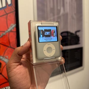 Könnte beinhalten: Silberner iPod Classic in einer transparenten Kunststoffhülle, der eine Musik-Playlist mit Albumcovern anzeigt. Der Bildschirm zeigt "Nascita Di Un Mito Maria Callas". Das Gerät hat ein kreisförmiges Steuerkreuz und eine Menütaste.