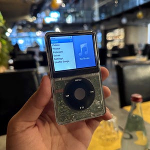 May include: A clear iPod music player with a black click wheel and a color screen displaying the menu options. The screen shows the words "Music", "Videos", "Photos", "Podcasts", "Extras", "Settings", and "Shuffle Songs". The screen also displays a musical note icon.