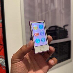 Könnte beinhalten: Ein kleines, lila-weißes iPod Nano wird in einer Hand gehalten. Der Bildschirm zeigt farbige Symbole für Musik, Videos, Fitness, Podcasts, Fotos und Radio an. Das Gerät hat einen weißen Bildschirm und eine kreisförmige Taste darunter.