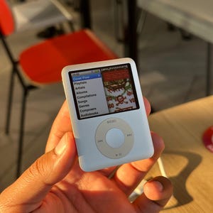 Könnte beinhalten: Ein weißer iPod-Musikplayer mit einem Bildschirm, der ein Wiedergabelistenmenü anzeigt. Der Bildschirm zeigt die Uhrzeit 12:55 und den Text "Cover Flow". Das Gerät verfügt über ein kreisförmiges Bedienfeld mit einer Menütaste und Navigationselementen.