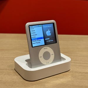 Könnte beinhalten: Silberner iPod Digital-Media-Player in einer weißen Dockingstation. Der Bildschirm zeigt das iPod-Menü mit Musik, Videos und Einstellungen. Das Apple-Logo ist sichtbar. Die Dockingstation ist weiß und rechteckig.