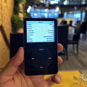 Könnte beinhalten: Ein schwarzer iPod-Digital-Media-Player mit einem kreisförmigen Bedienfeld und einem Bildschirm, der Geräteinformationen anzeigt. Der Bildschirm zeigt Details wie Songanzahl, Speicherkapazität und Softwareversion an. Das Gerät wird in einer Hand gehalten.