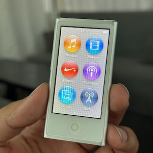 Könnte beinhalten: Ein silberner iPod Nano mit Touchscreen, der App-Symbole für Musik, Videos, Fitness, Podcasts, Fotos und Radio anzeigt. Die Uhrzeit 14:11 wird angezeigt. Das Gerät wird in einer Hand gehalten.