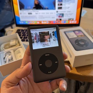 Könnte beinhalten: Ein dunkelgrauer iPod Classic mit schwarzem Bedienfeld und einem Bildschirm, der Albumcover anzeigt. Das Gerät wird in einer Hand gehalten, mit einem Laptop und Zubehör im Hintergrund. Der Bildschirm zeigt den Text "Bugiardo Fabri Fibra".
