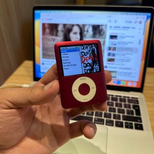Könnte beinhalten: Ein roter iPod Nano wird vor einem Laptop gehalten. Der iPod-Bildschirm zeigt ein Menü und die Worte "DON'T DEAL DON'T" im Graffiti-Stil. Der Laptop-Bildschirm zeigt ein Video. Der iPod hat ein kreisförmiges Bedienfeld.