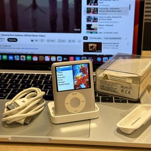 Könnte beinhalten: Ein silberner iPod Classic mit weißer Dockingstation, der ein farbenfrohes Bild auf seinem Bildschirm anzeigt. Ein weißes USB-Kabel und eine transparente Kunststoffhülle sind ebenfalls sichtbar. Der Bildschirm zeigt die Menüoptionen: Musik, Videos, Fotos, Podcasts, Extras, Einstellungen und Zufallswiedergabe.
