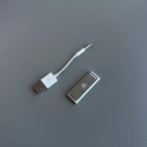May include: A white USB to 3.5mm audio adapter cable and a silver iPod Shuffle. The cable has a USB-A connector on one end and a 3.5mm audio jack on the other. The iPod Shuffle has the Apple logo.