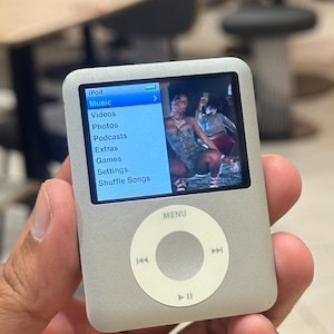 Könnte beinhalten: Ein silberner iPod Digital-Media-Player mit einem Bildschirm, der ein Musikvideo anzeigt. Der Bildschirm zeigt die Menüoptionen: Musik, Videos, Fotos, Podcasts, Extras, Spiele, Einstellungen und Zufallswiedergabe. Der iPod hat ein kreisförmiges Bedienfeld.