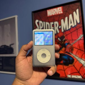 Może przedstawiać: Srebrny iPod Classic trzymany w dłoni, wyświetlający menu muzyczne na ekranie. Urządzenie posiada okrągły panel sterowania i ekran. Plakat Marvel Spider-Man znajduje się w tle.