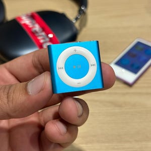 Pode incluir: Um pequeno reprodutor de música digital quadrado azul claro com um painel de controle circular branco. O painel de controle tem um botão central de reprodução/pausa e botões de navegação circundantes. Um iPod branco é visível no fundo.
