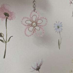 Könnte beinhalten: Ein rosa Perlenblumen-Schlüsselanhänger mit Perlenmitte und goldenen Drahtakzenten. Die Blume ist an einer silbernen Kette und einem Schlüsselring befestigt. Der Hintergrund zeigt Aquarell-Blumendesigns.