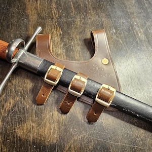 May include: A medieval sword with a wooden handle and a silver crossguard, resting in a black sheath. The sheath is secured by three brown leather straps with gold-colored buckles. A brown leather scabbard with a fleur-de-lis design is visible.