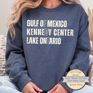 May include: A dark blue heathered crewneck sweatshirt featuring white distressed text that reads GULF OF MEXICO, KENNEDY CENTER, and LAKE ONTARIO. The letters F, D, and T are highlighted in a different font style within the words.
