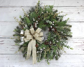 May include: A festive Christmas wreath with a mix of evergreen branches, silver berries, and pinecones. A large, beige bow with silver ribbon accents the wreath. The wreath is displayed against a white, rustic wooden background.