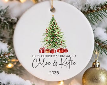 May include: White ceramic Christmas ornament with a watercolor Christmas tree design, red gift boxes, and the text "FIRST CHRISTMAS ENGAGED Chloe & Katie 2025". The ornament hangs from a gold chain.