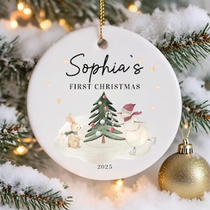 May include: A white ceramic Christmas ornament with the text "Sophia's FIRST CHRISTMAS" and the year 2025. The ornament features a watercolor illustration of a snowman, a rabbit, and a Christmas tree. The ornament is hanging from a gold chain.