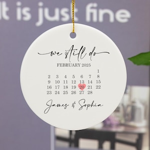 Könnte beinhalten: Weißer Keramik-Anhänger mit einem Kalender für Februar 2025, mit einem Herz auf dem 20. Der Text "we still do" ist in Kursivschrift über dem Kalender geschrieben. Die Namen "James & Sophia" stehen unter dem Kalender.