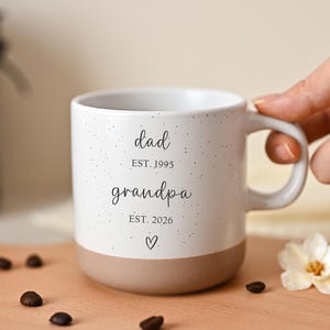 Peut inclure: Tasse en céramique blanche avec un motif moucheté et une base beige. La tasse porte l'inscription "dad EST. 1995 grandpa EST. 2026" et un symbole de cœur. La tasse est tenue par une main.