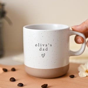 Op de afbeelding: Witte keramische mok met een gespikkeld ontwerp en een bruine basis. De mok heeft de tekst "oliva's dad" en een hartsymbool. De mok staat op een houten snijplank met koffiebonen en een witte bloem.