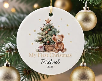Puede incluir: Un adorno redondo de cerámica blanca con una ilustración en acuarela de un árbol de Navidad y un osito de peluche. El texto dice My First Christmas, Michael y 2026. Cuelga de un cordón dorado sobre ramas de pino de color verde.