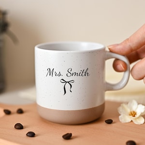 Puede incluir: Una taza de cerámica blanca con acabado moteado y base de color tostado sólido. El frente presenta el texto negro Mrs. Smith escrito en cursiva sobre un pequeño gráfico de lazo negro. La taza tiene un asa curva en el lado derecho.