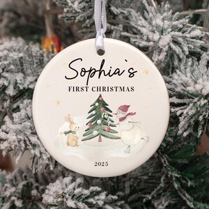 May include: A round Christmas ornament with the text "Sophia's First Christmas" and the year 2025. The ornament features a watercolor illustration of a snowman, a bunny, and a Christmas tree. The ornament hangs from a white ribbon.